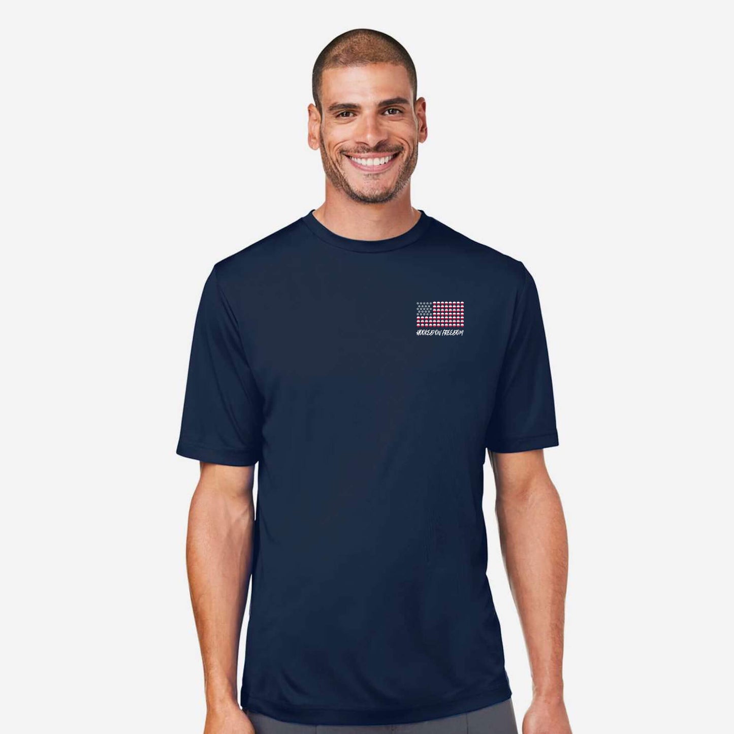 A smiling man wearing a navy blue crewneck t-shirt with a small red, white, and blue grid graphic on the left chest, set against a plain light gray background.