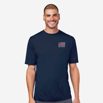 A smiling man wearing a navy blue crewneck t-shirt with a small red, white, and blue grid graphic on the left chest, set against a plain light gray background.
