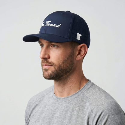 A man wearing a navy blue baseball cap with white embroidered text reading 'The Thousand' and a white Minnesota logo, paired with a gray t-shirt against a light gray background.