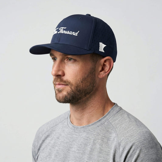 A man wearing a navy blue baseball cap with white embroidered text reading 'The Thousand' and a white Minnesota logo, paired with a gray t-shirt against a light gray background.